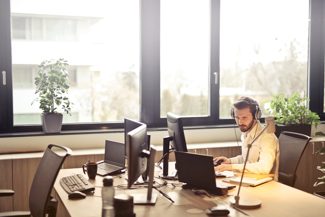 heros-img A businessman sits at a desk using multiple computers and a headset in a well-lit modern office.